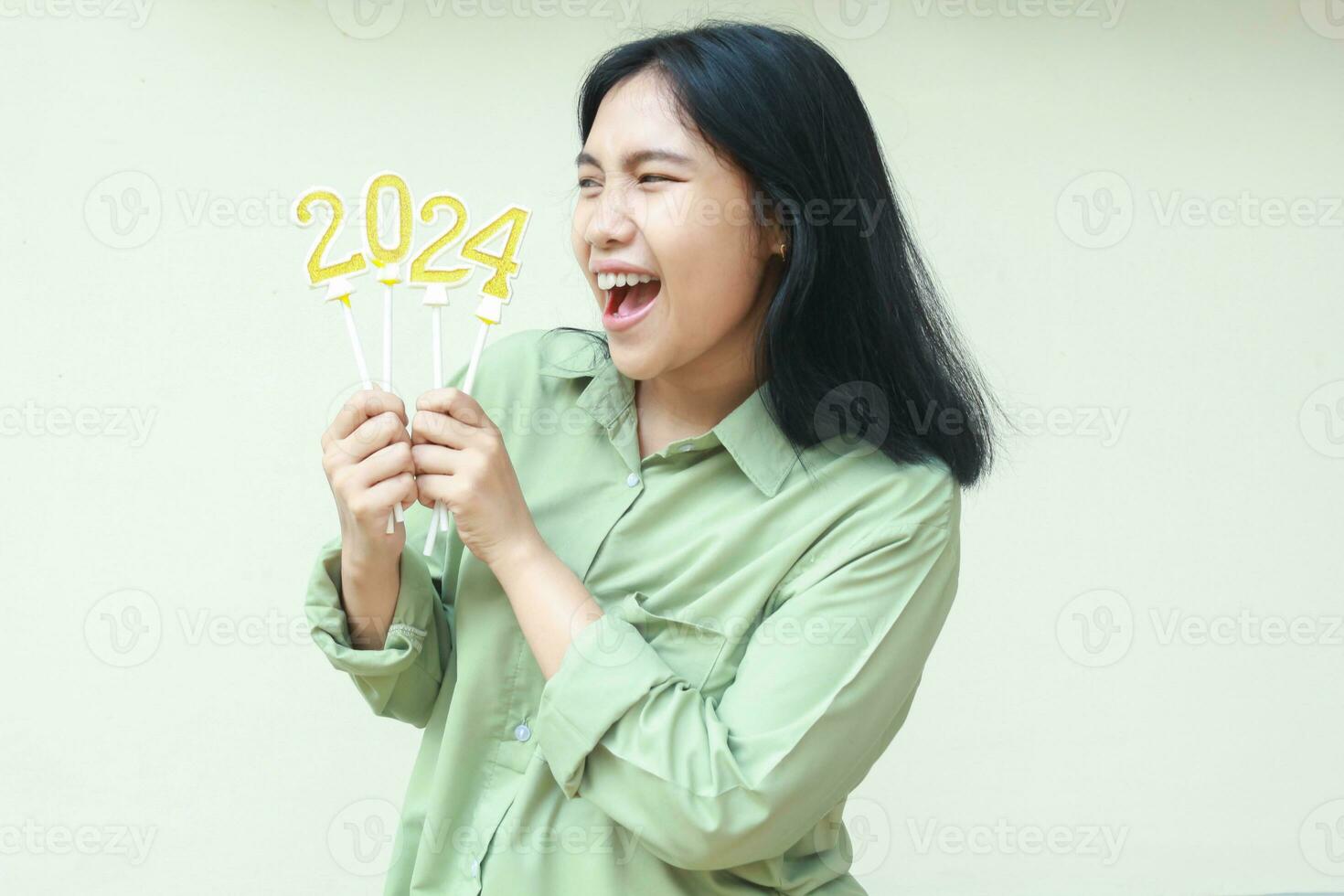 conmocionado asiático joven mujer hipster gritando mientras mirando aparte a 2024 número vela elevado en mano sostener vistiendo verde terminado Talla camisa casual, aislado terminado blanco, nuevo años víspera concepto foto