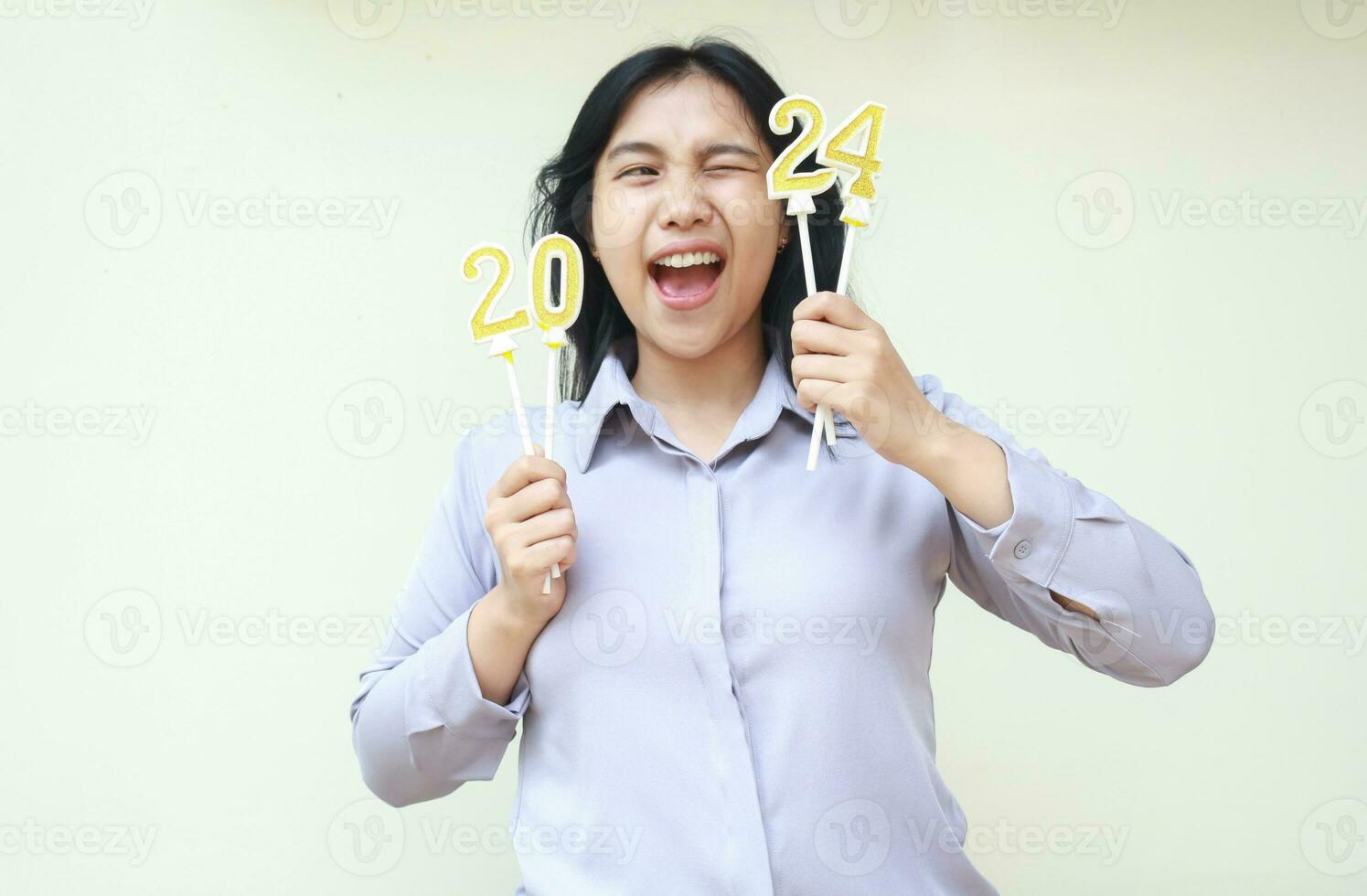 emocionado asiático joven mujer levantamiento 2024 figura vela participación en mano, hembra compañero de trabajo sonriente con abierto boca y mirando aparte a número 24 vestir gris formal traje, aislado en blanco foto