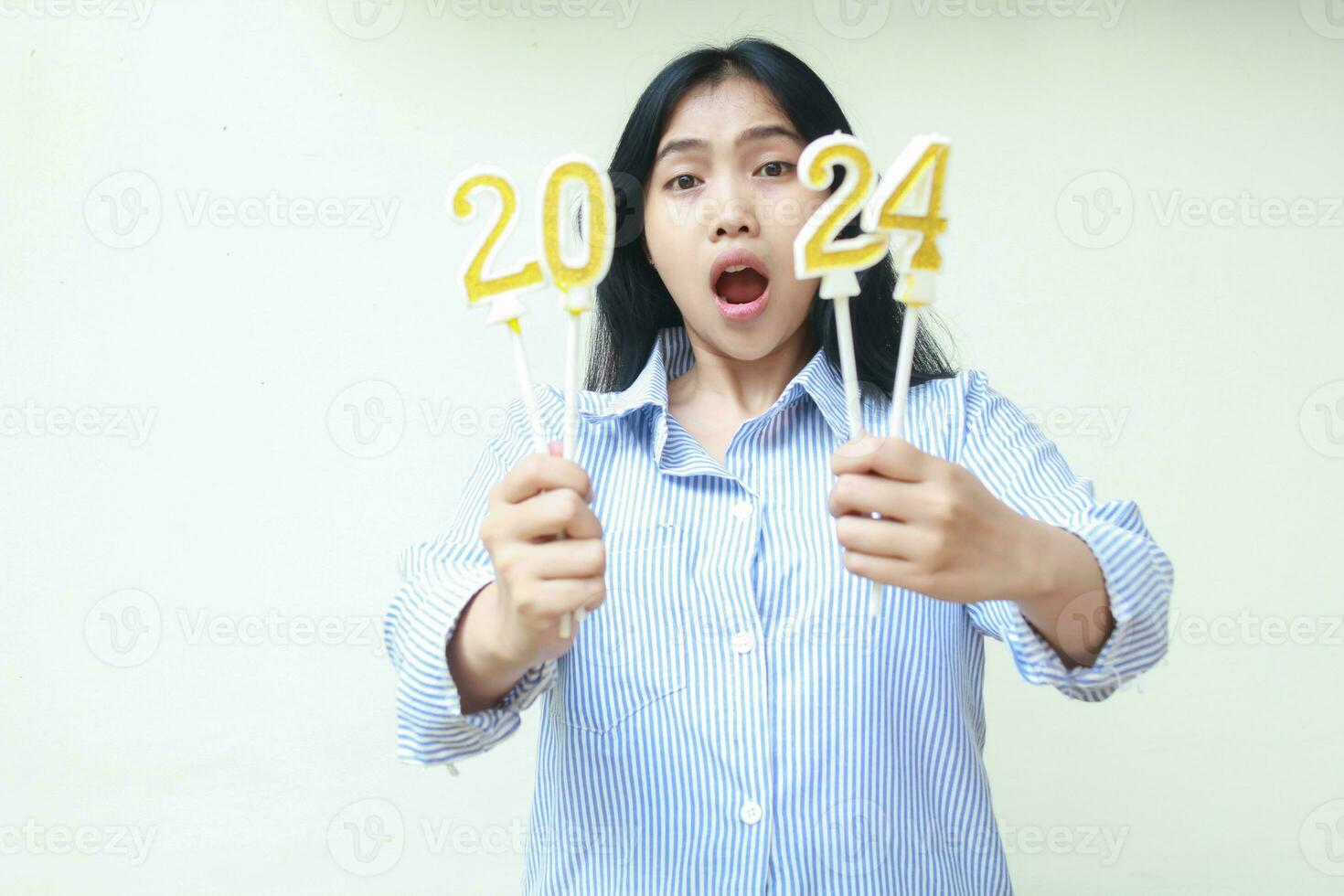 conmocionado asiático joven mujer aturdido con abierto boca mientras demostración 2024 número vela participación en mano, hembra compañero de trabajo vistiendo azul rayas camisa casual, nuevo años víspera concepto, aislado en blanco foto