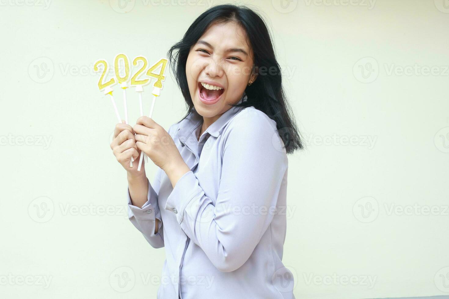 emocionado joven negocio mujer asiático gritando con abierto boca a cámara con levantamiento brazo participación 2024 número vela, extático hembra compañero de trabajo vistiendo gris formal traje a celebrando nuevo años víspera foto