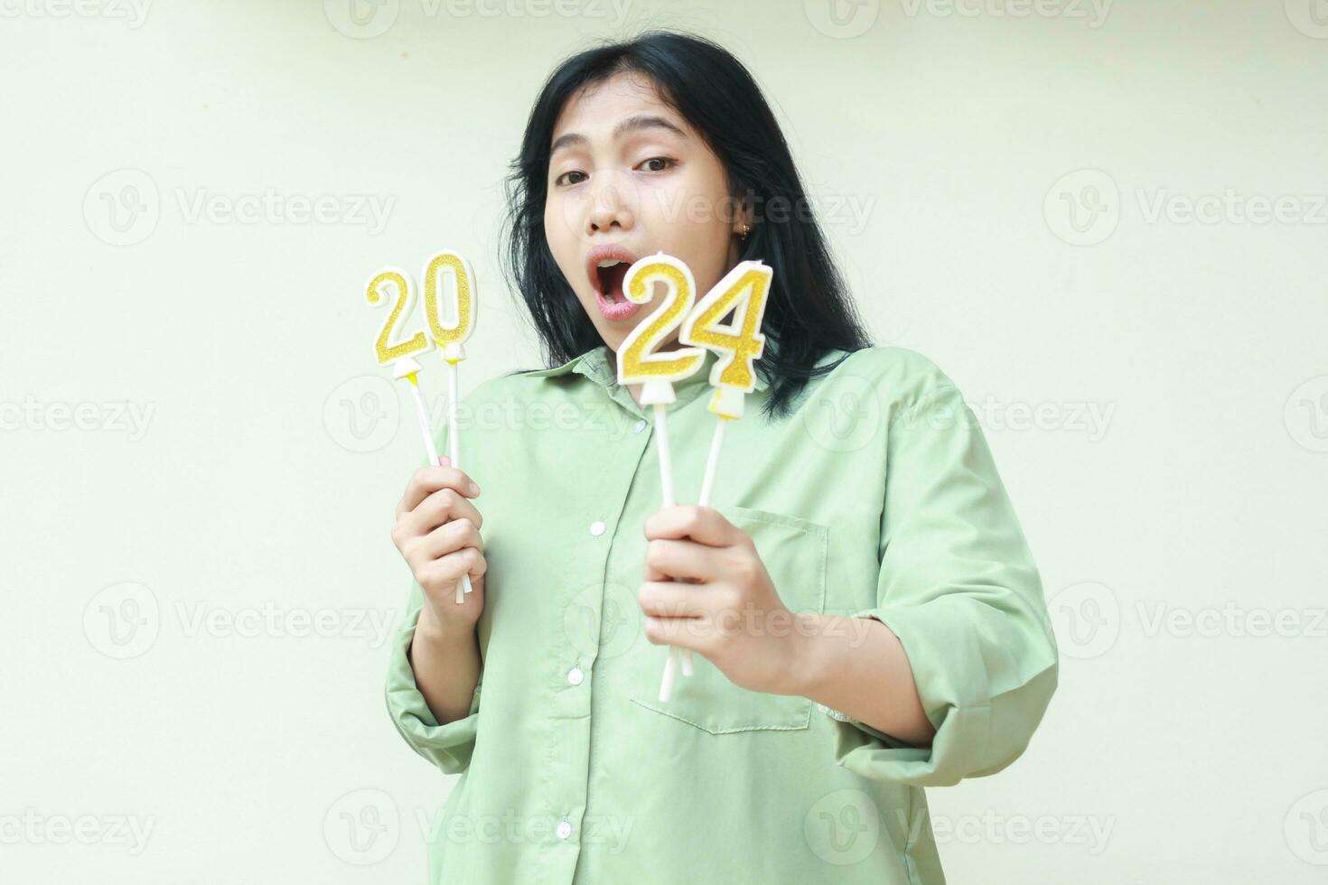 asian young woman surprised with open mouth wearing green over size shirt with holding 2024 number candle, looking to camera, isolated over white background photo