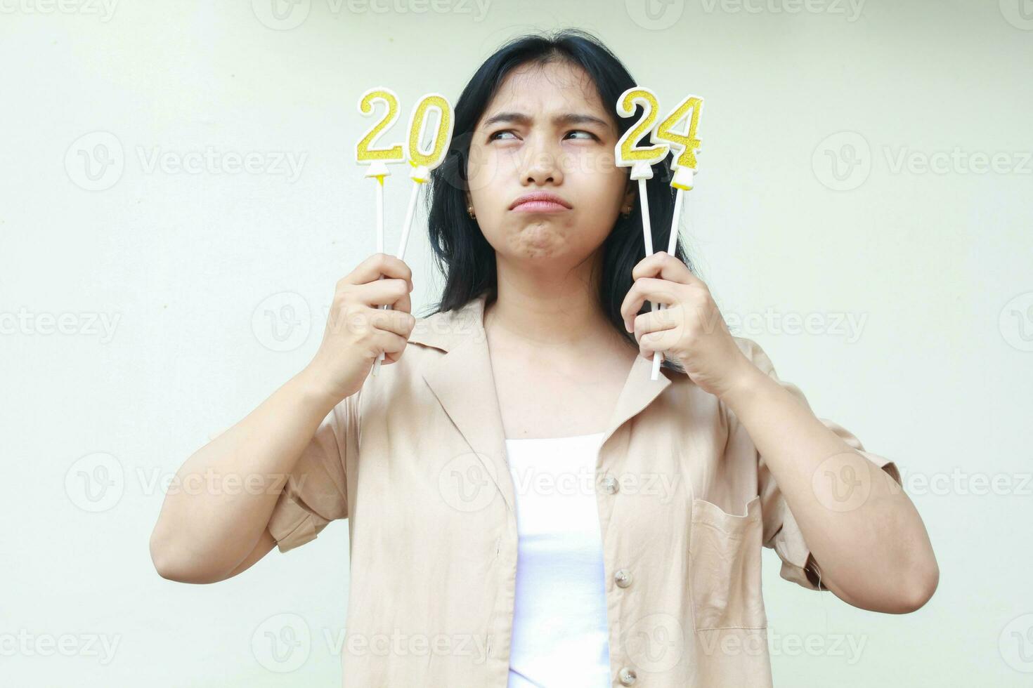 thoughtful asian young woman looking aside to number 20 of 2024 figure candle holding on hand wearing stylish brown outer, isolated on white background, new years eve concept photo