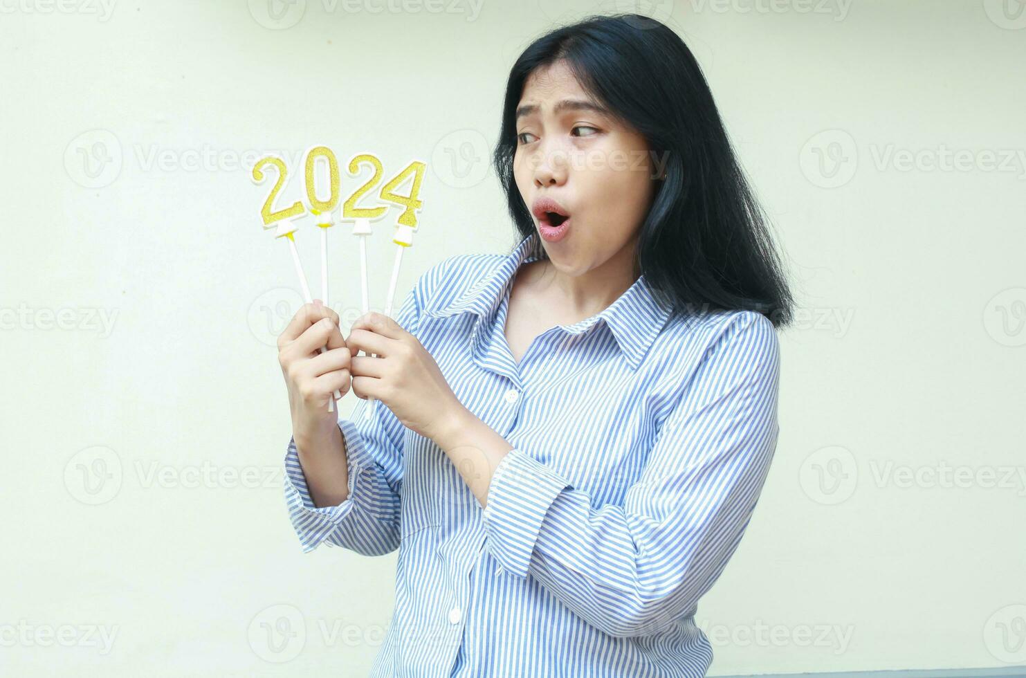 joven negocio mujer asiático aturdido y mirando aparte a vela número 2024, hembra compañero de trabajo vistiendo azul rayas casual camisa, aislado en blanco antecedentes foto