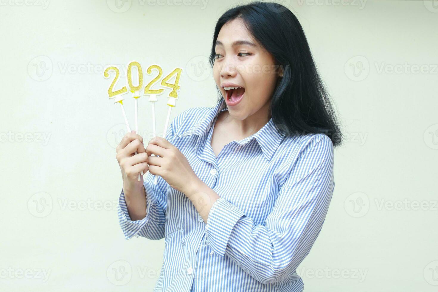 sorprendido asiático joven mujer gritando y mirando aparte a 2024 número velas sostener en mano vistiendo casual azul rayas camisa, aislado en blanco, nuevo años víspera celebracion foto