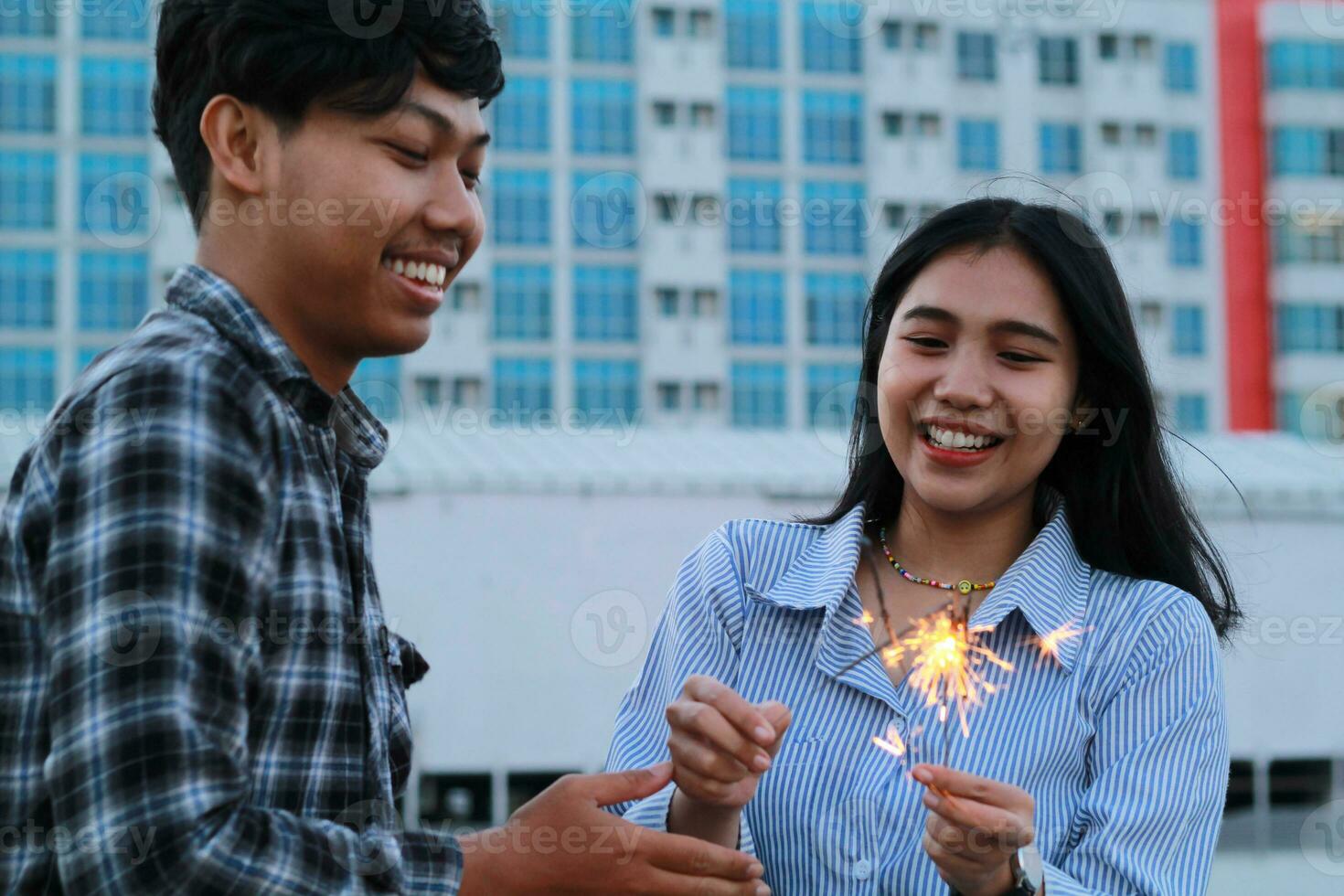 playful asian young couple burning sparkler with smiling face for new year celebration, on urban city building background wearing flannel and stripes shirt casual photo
