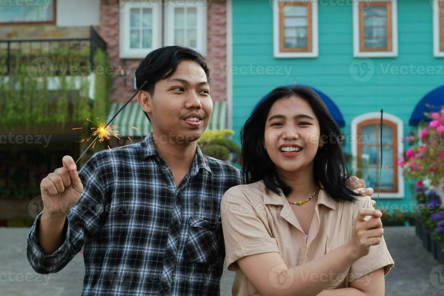 candid asian couple enjoying new year celebration by holding sparkler firework in the house yard green garden wearing casual outer and flannel photo