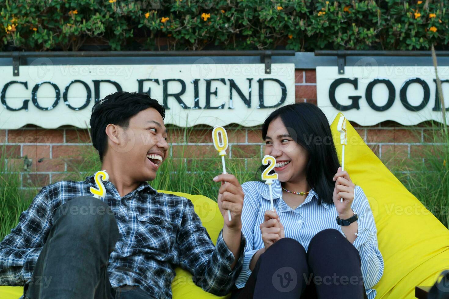 happy asian couple laughing looking to each other with raised hand holding candle number 20 24 sitting on the yellow couch or bean bag in the house backyard with brick wall and flower grass background photo