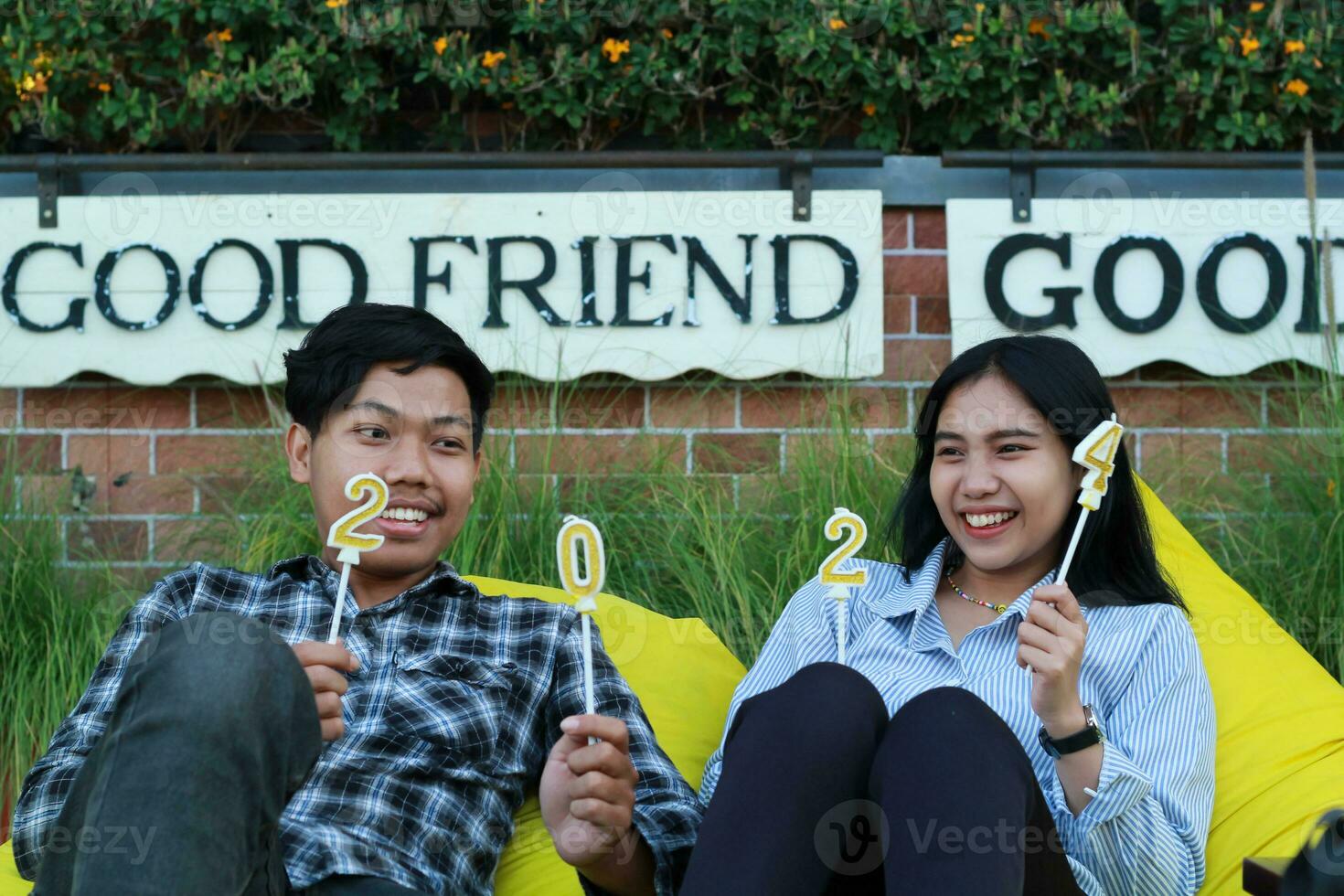 teenager couple sitting on the yellow couch with happy expression while holding golden candle number 20 24, man and woman hipster celebrate new year in the house yard with brick wall background photo