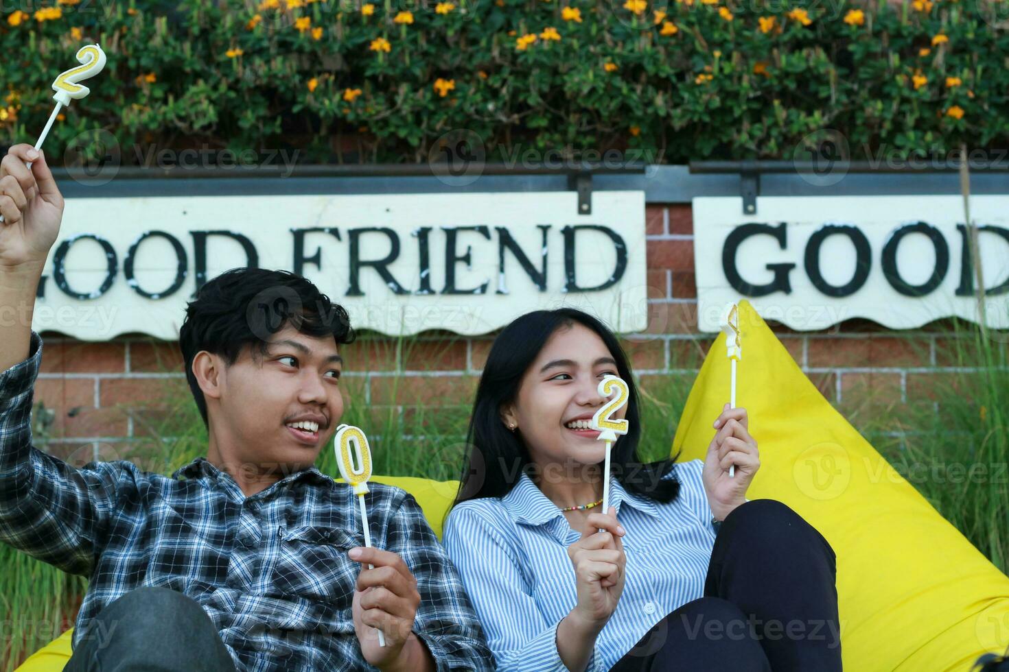 happy multiracial couple looking away with raising hand holding 20 24 candle number, sitting on the yellow bean bag couch with brick wall and flower background photo