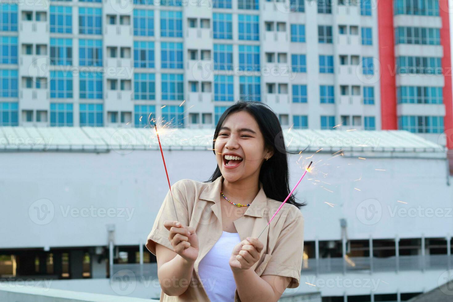 excited asian young woman holding sparkler to celebrating new year eve in rooftop apartment with city building background photo