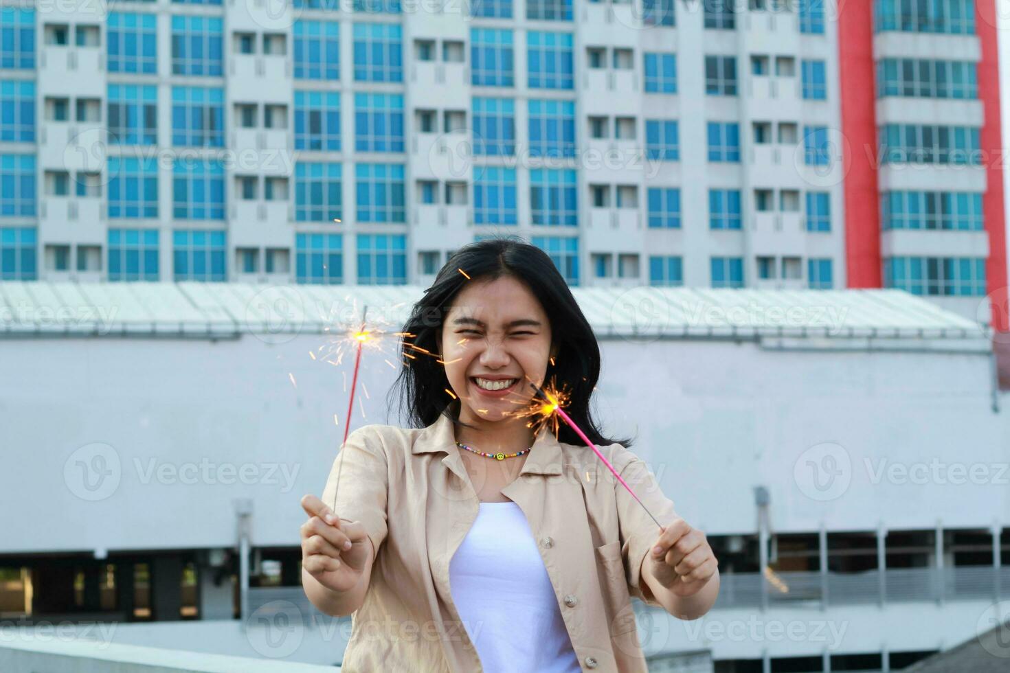 excited asian young woman holding sparkler to celebrating new year eve in rooftop apartment with city building background photo