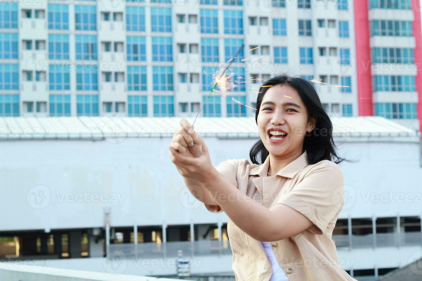 beautiful asian young woman holding sparklers firework and laughing in new year eve celebration standing in roof top outdoor with urban building background photo