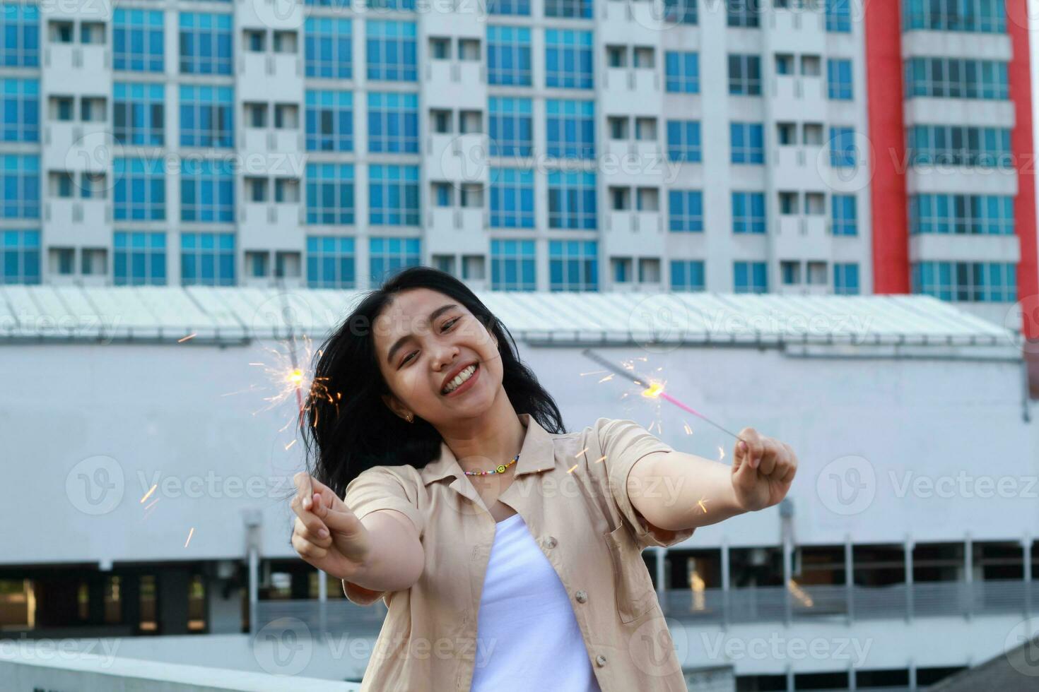 carefree asian young woman holding sparkler celebrate new year eve with laugh and dance in rooftop apartment outdoor with city building background photo