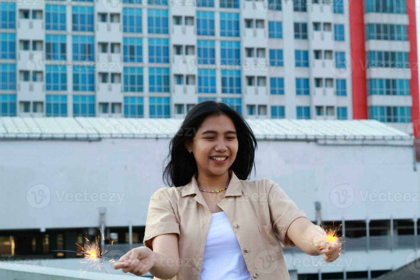 carefree asian young woman holding sparkler celebrate new year eve with laugh and dance in rooftop apartment outdoor with city building background photo