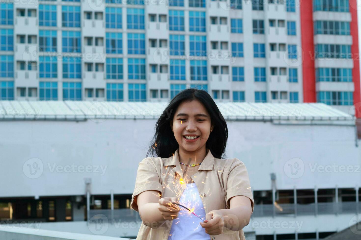 carefree asian young woman holding sparkler celebrate new year eve with laugh and dance in rooftop apartment outdoor with city building background photo