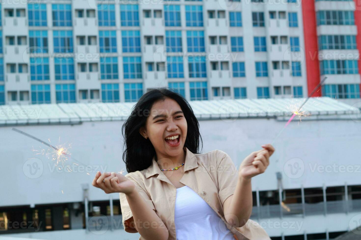 carefree asian young woman holding sparkler celebrate new year eve with laugh and dance in rooftop apartment outdoor with city building background photo