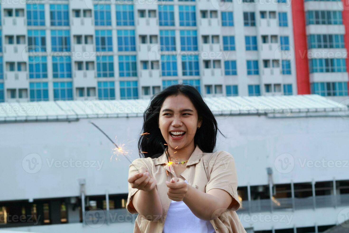 carefree asian young woman holding sparkler celebrate new year eve with laugh and dance in rooftop apartment outdoor with city building background photo