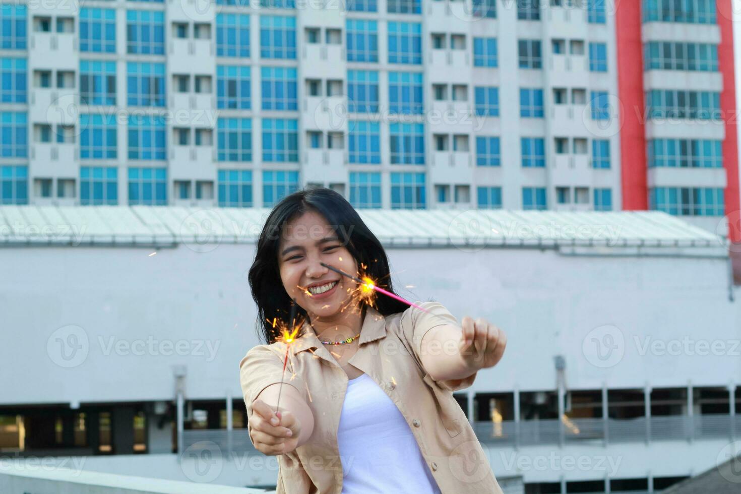 contento asiático joven mujer hipster participación bengalas Fuegos artificiales celebrar nuevo año víspera con bailando en techo parte superior Departamento con urbano edificio antecedentes foto