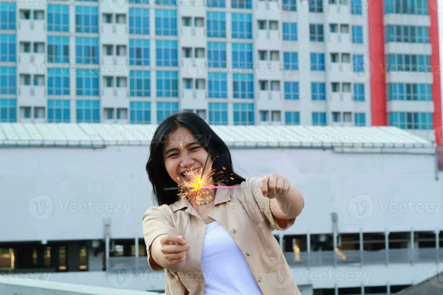 contento asiático joven mujer hipster participación bengalas Fuegos artificiales celebrar nuevo año víspera con bailando en techo parte superior Departamento con urbano edificio antecedentes foto