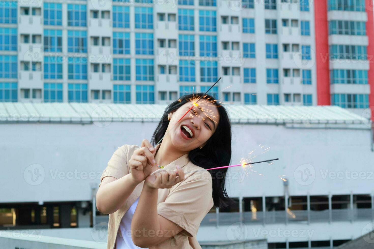 contento asiático joven mujer hipster participación bengalas Fuegos artificiales celebrar nuevo año víspera con bailando en techo parte superior Departamento con urbano edificio antecedentes foto