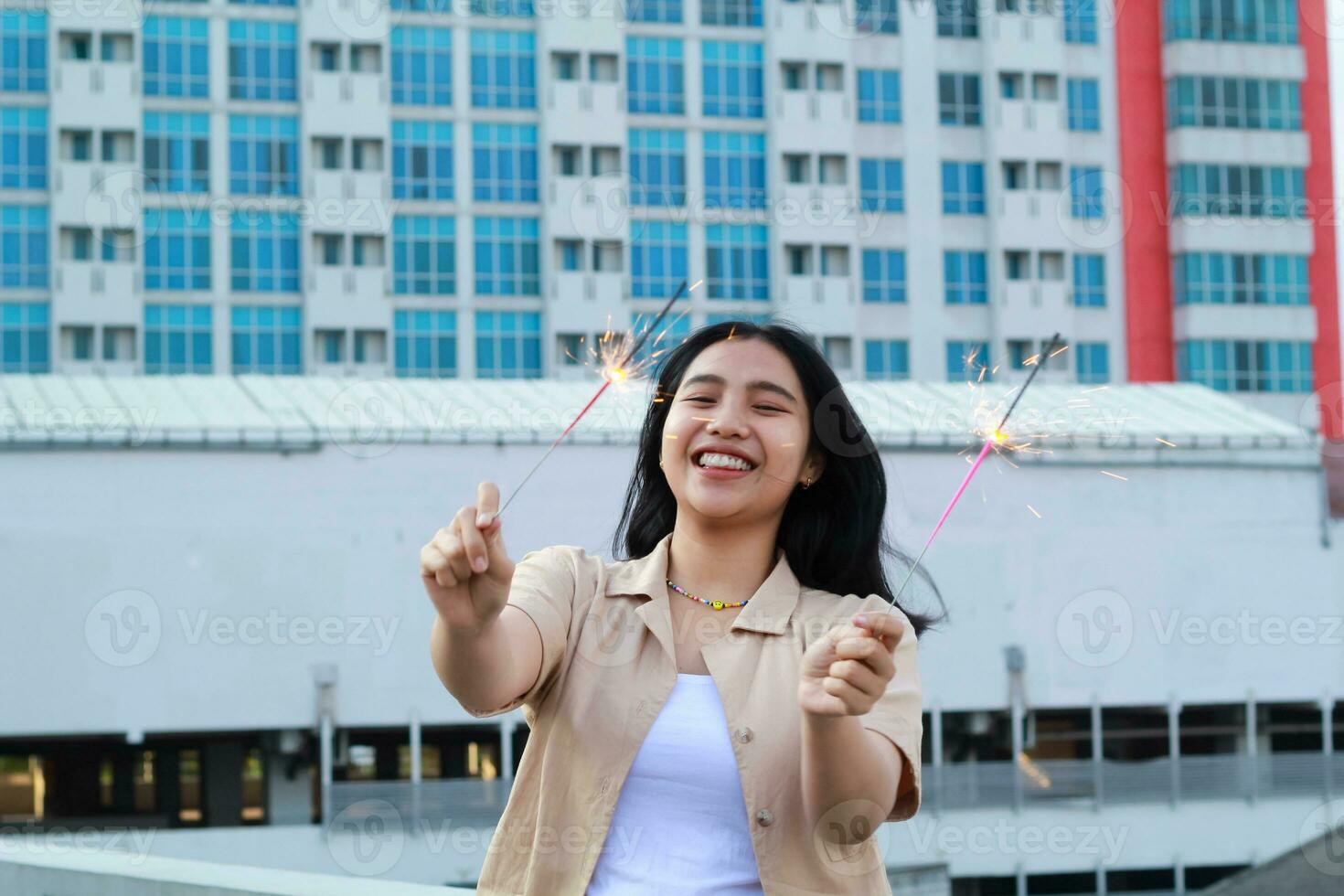 contento asiático joven mujer hipster participación bengalas Fuegos artificiales celebrar nuevo año víspera con bailando en techo parte superior Departamento con urbano edificio antecedentes foto