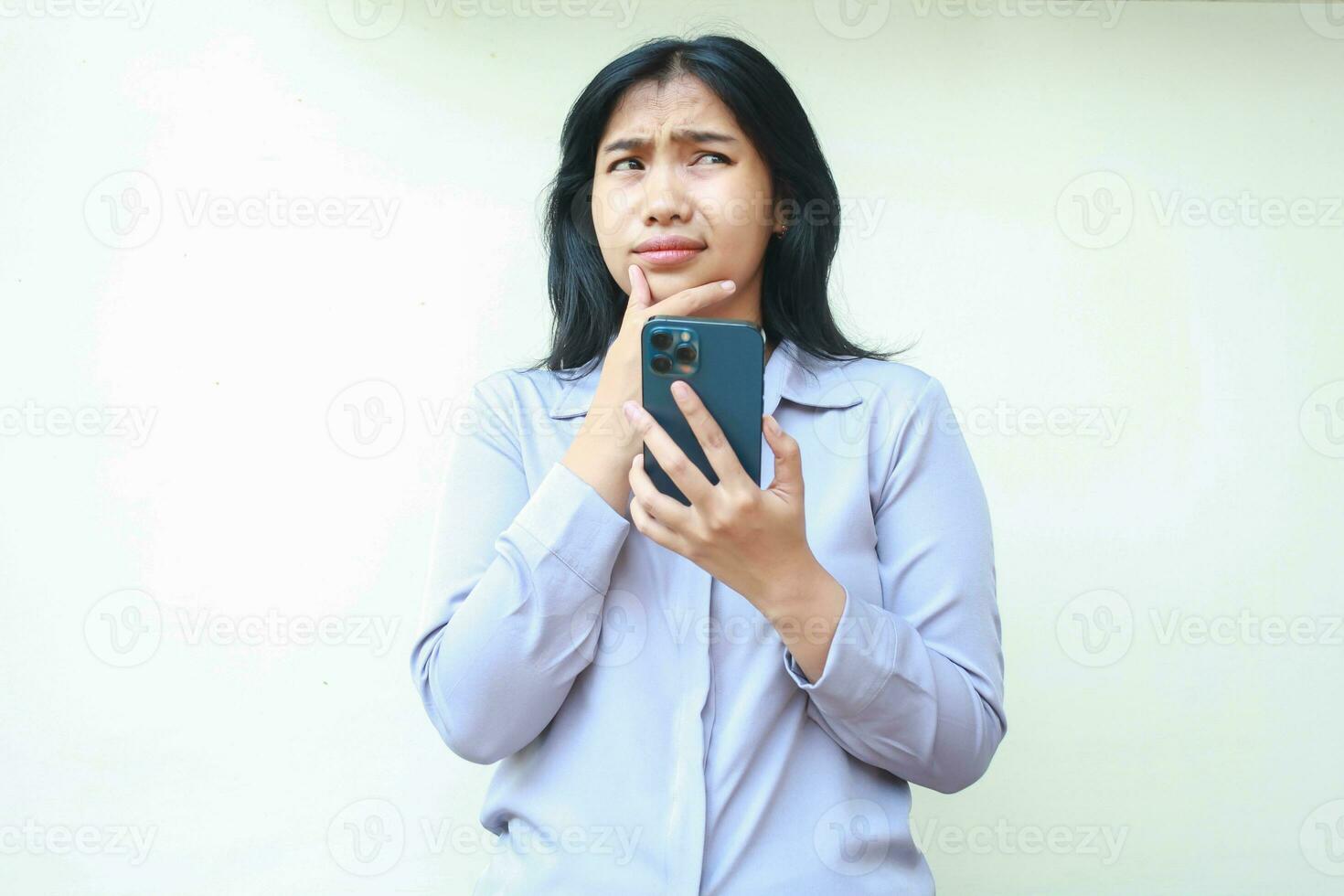 portrait of confused asian young woman using smartphone thinking seriously with hand on chin looking away, beautiful female considering choices wearing formal suit isolated in white background photo