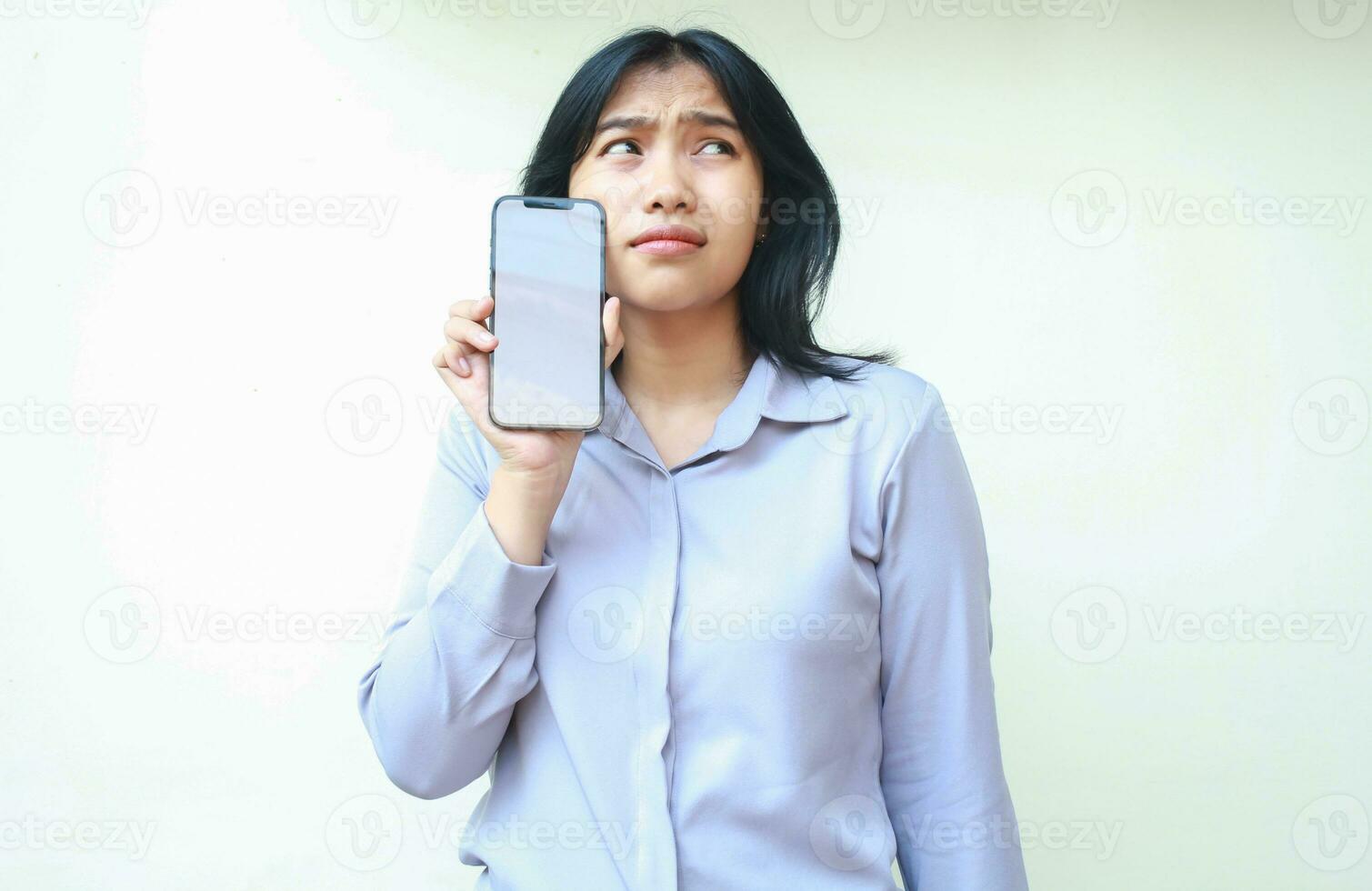 unhappy sad asian young woman showing white screen of smartphone and looking above thinking, female wear formal suit isolated in white background, mockup photo