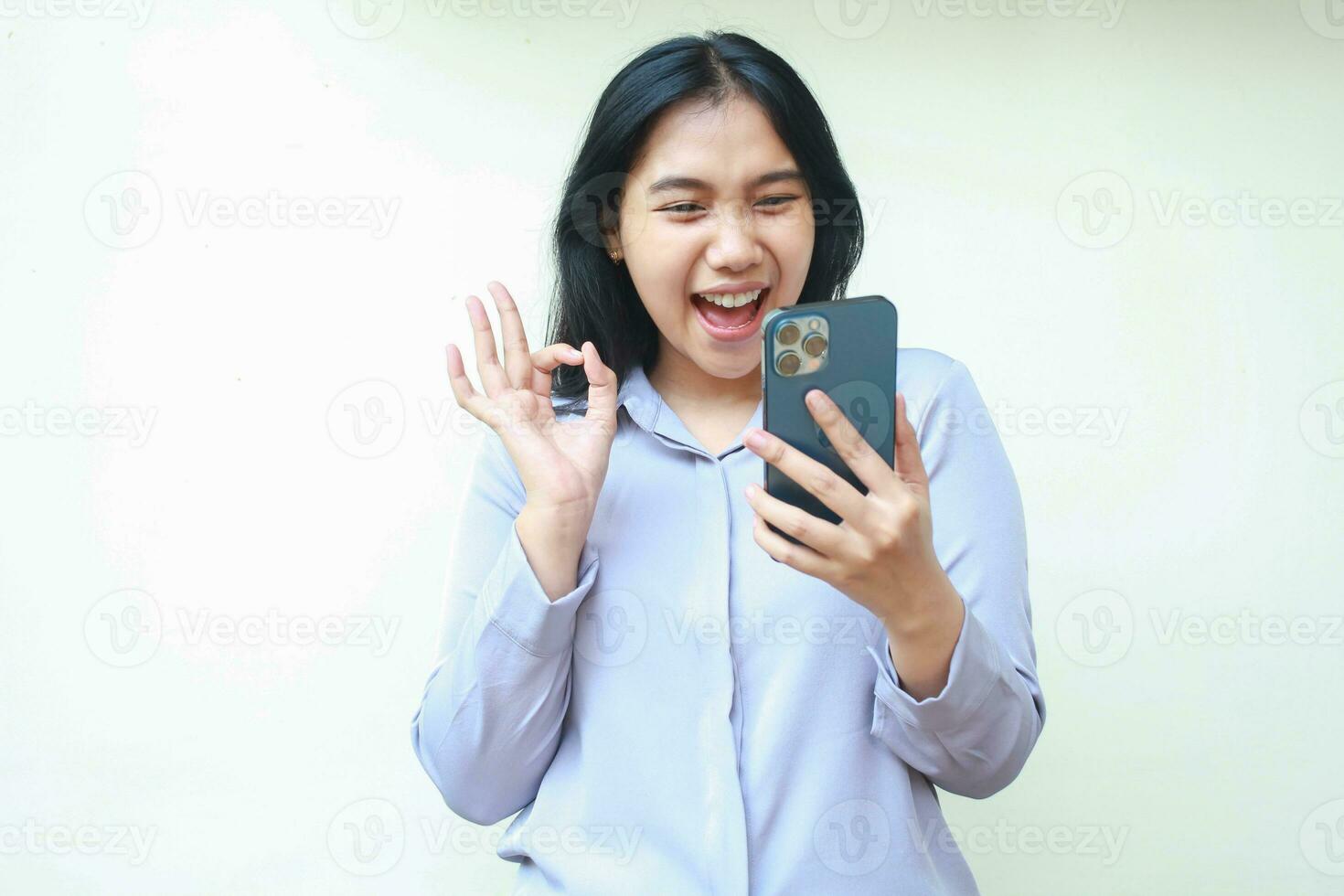 excited carefree asin young business woman holding mobile phone and gesturing ok sign while video calling, smiling female with approval pose wearing formal shirt isolated in white background photo