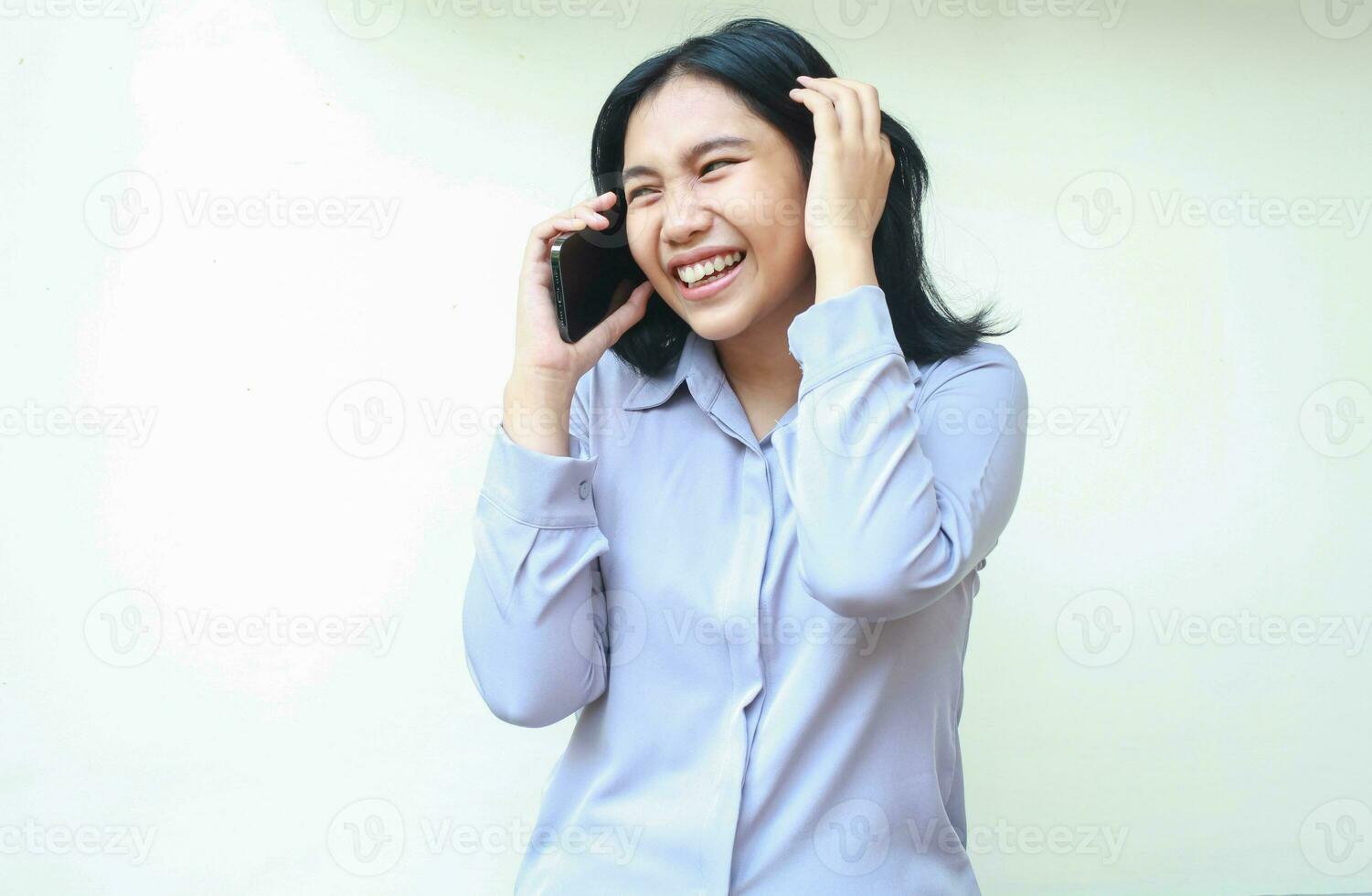 Encantado asiático joven negocio mujer riendo mientras hablar en teléfono inteligente participación su oscuro pelo Mira lejos vestir formal púrpura camisa en pie terminado blanco antecedentes foto