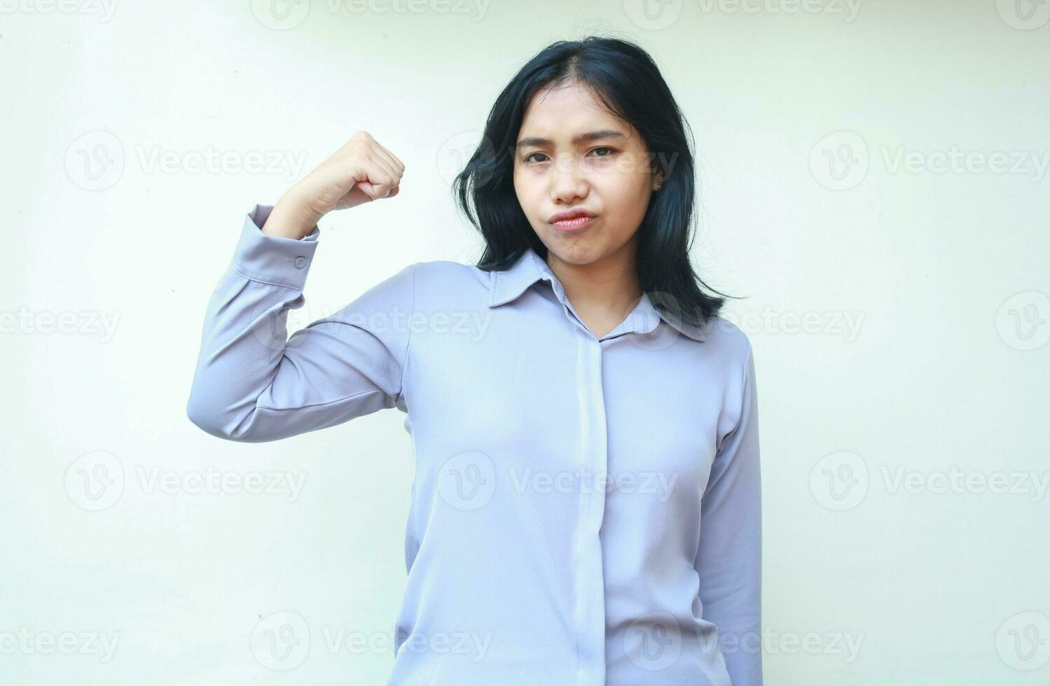 fuerte asiático negocio mujer levantamiento brazos demostración bíceps músculo fuerza con grave cara expresión mirando a cámara vistiendo formal camisa en pie terminado blanco antecedentes foto