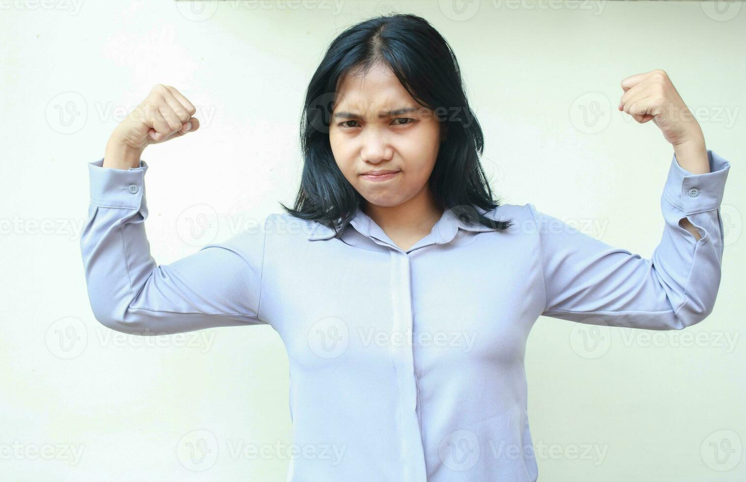 fuerte asiático negocio mujer levantamiento brazos demostración bíceps músculo fuerza con grave cara expresión mirando a cámara vistiendo formal camisa en pie terminado blanco antecedentes foto