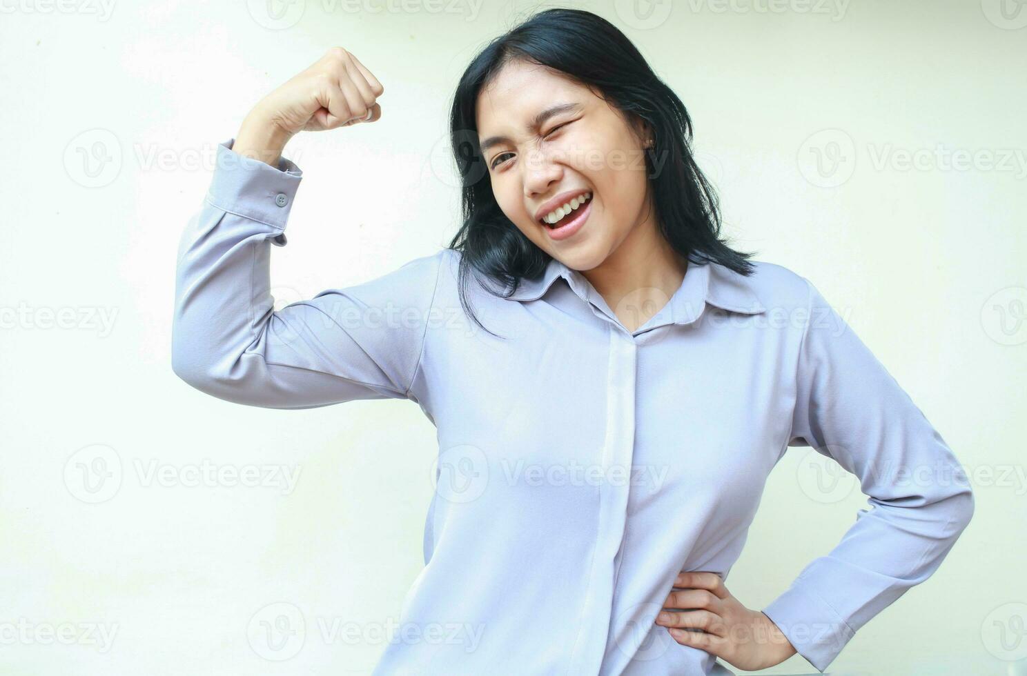 carefree powerful asian young business woman raising arm excited showing biceps strenght with hands on waist, blink eye flirting, wearing formal shirt standing over white background, looking at camera photo