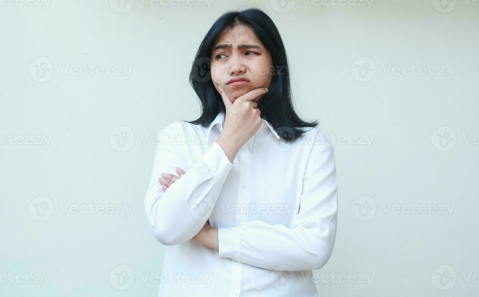 studio shot of worried young asian business woman folded arms looking aside with hand on chin wearing white formal suit, negative afraid expression, standing photo