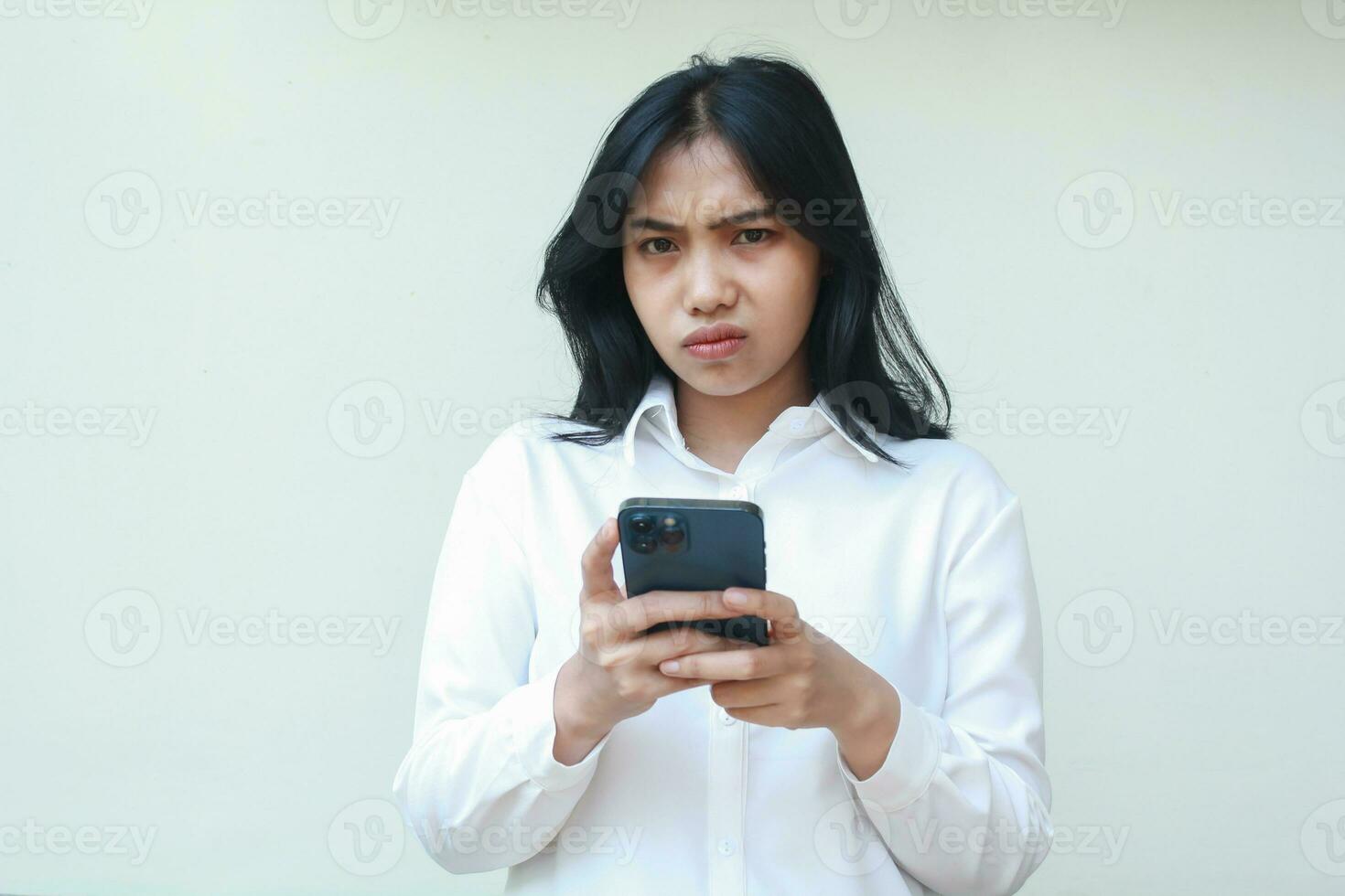 sad irritated asian business woman 20s glaring to camera holding mobile phone unhappy, dissatisfied and displeased facial expression, wearing white formal suit standing over isolated background photo