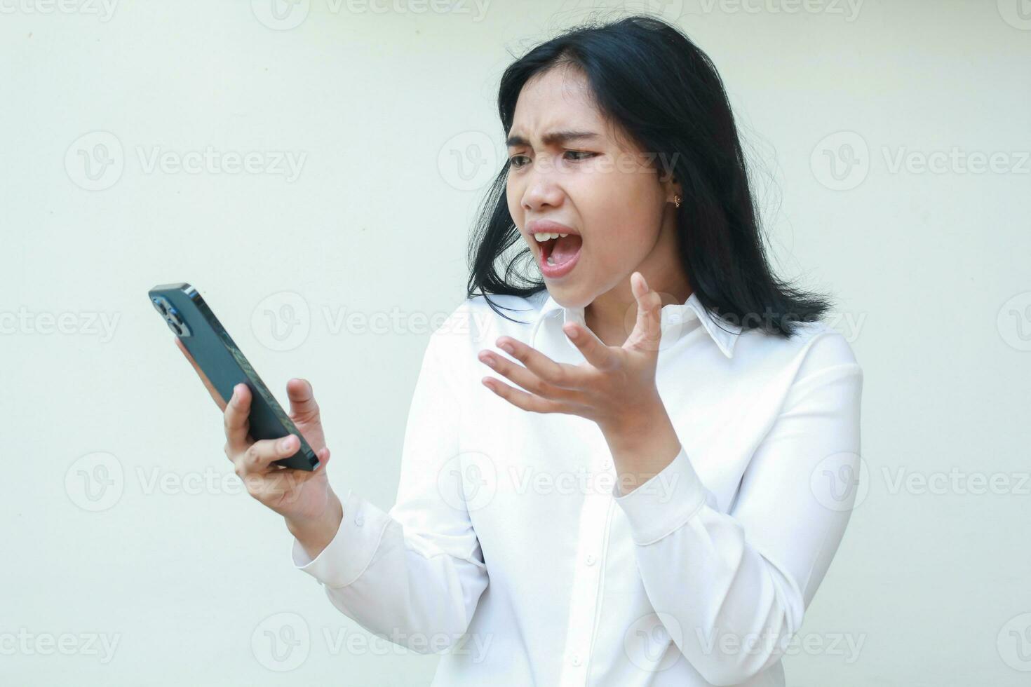 disgruntled furious asian woman looking to smartphone with screaming dissatisfied and raising palm, speaking and questioning gesture, wearing white formal suit standing over isolated background photo