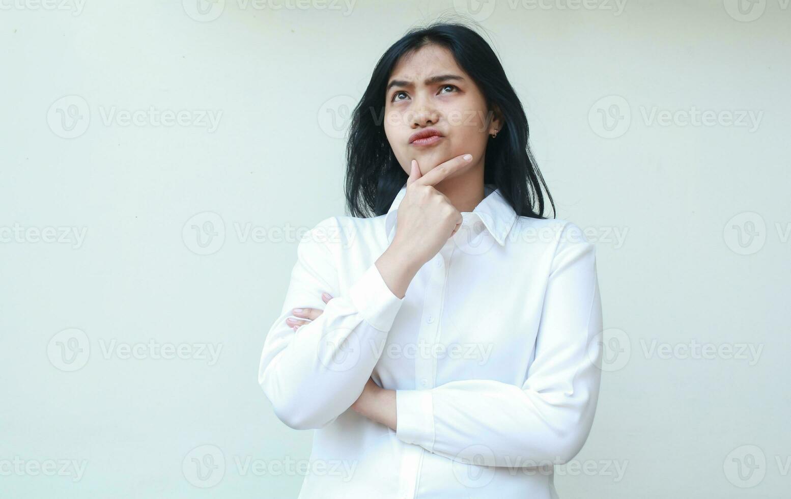 retrato de pensativo asiático joven negocio mujer mirando arriba suspicaz con manos en barbilla y brazos doblada vestir blanco formal camisa, en pie foto