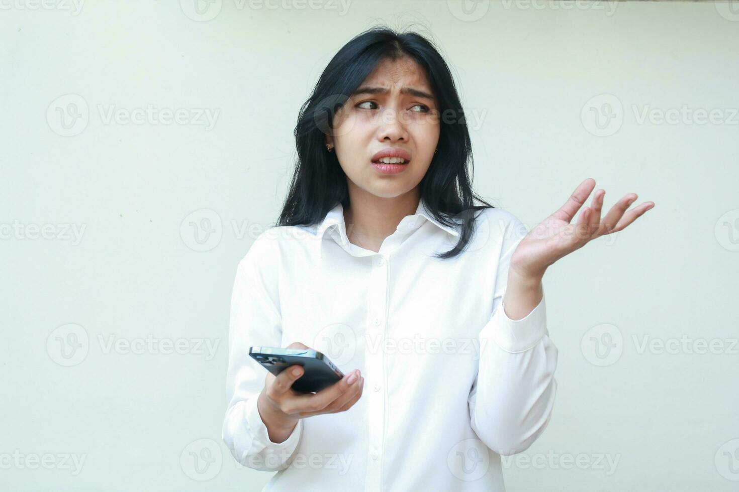 portrait of thoughtful asian business woman manager looking aside to empty space confused, raising palms showing presenting gesture, holding smartphone wearing white formal shirt, standing photo