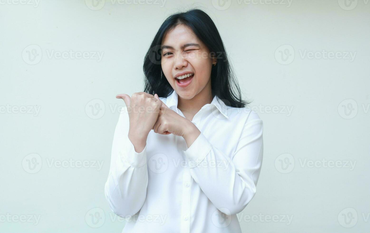 studio shot of excited asian young business woman pointing to side empty space with her thumbs look at camera flirting blink eye wearing white formal suit shirt isolated on white background photo