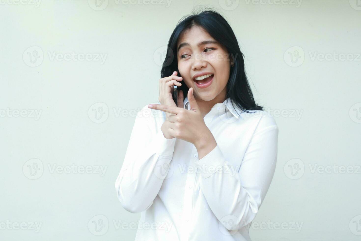retrato de éxito asiático joven negocio mujer gerente Hablando en teléfono inteligente con emoción Mira aparte señalando dedo junto a a vacío espacio con pulgares arriba vistiendo blanco formal traje camisa, en pie foto