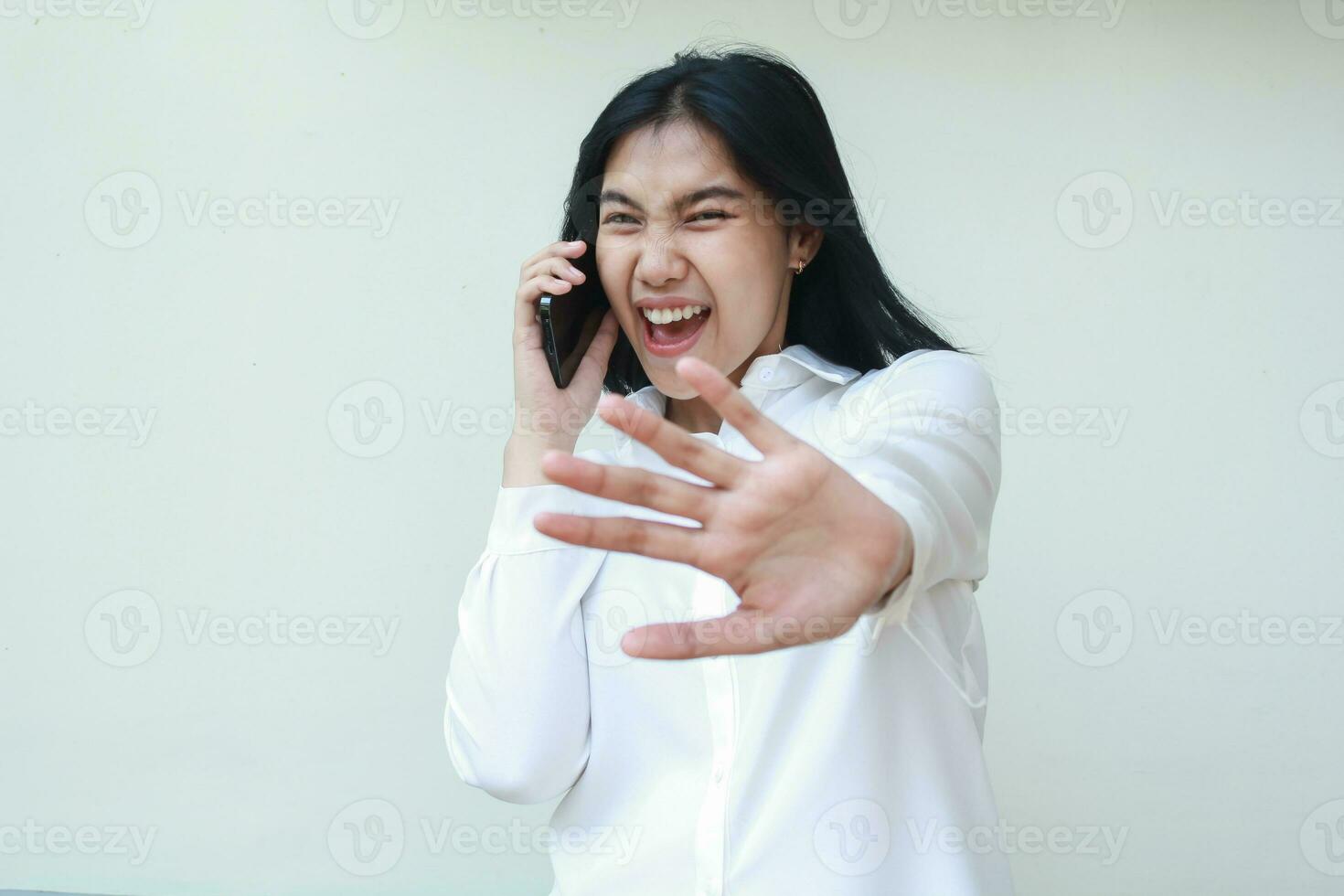 portrait of excited asian young woman secretary speaking on mobile phone reaching hands palm towards camera as stop signs wear white formal shirt, warning forbidden gesture, standing isolated photo