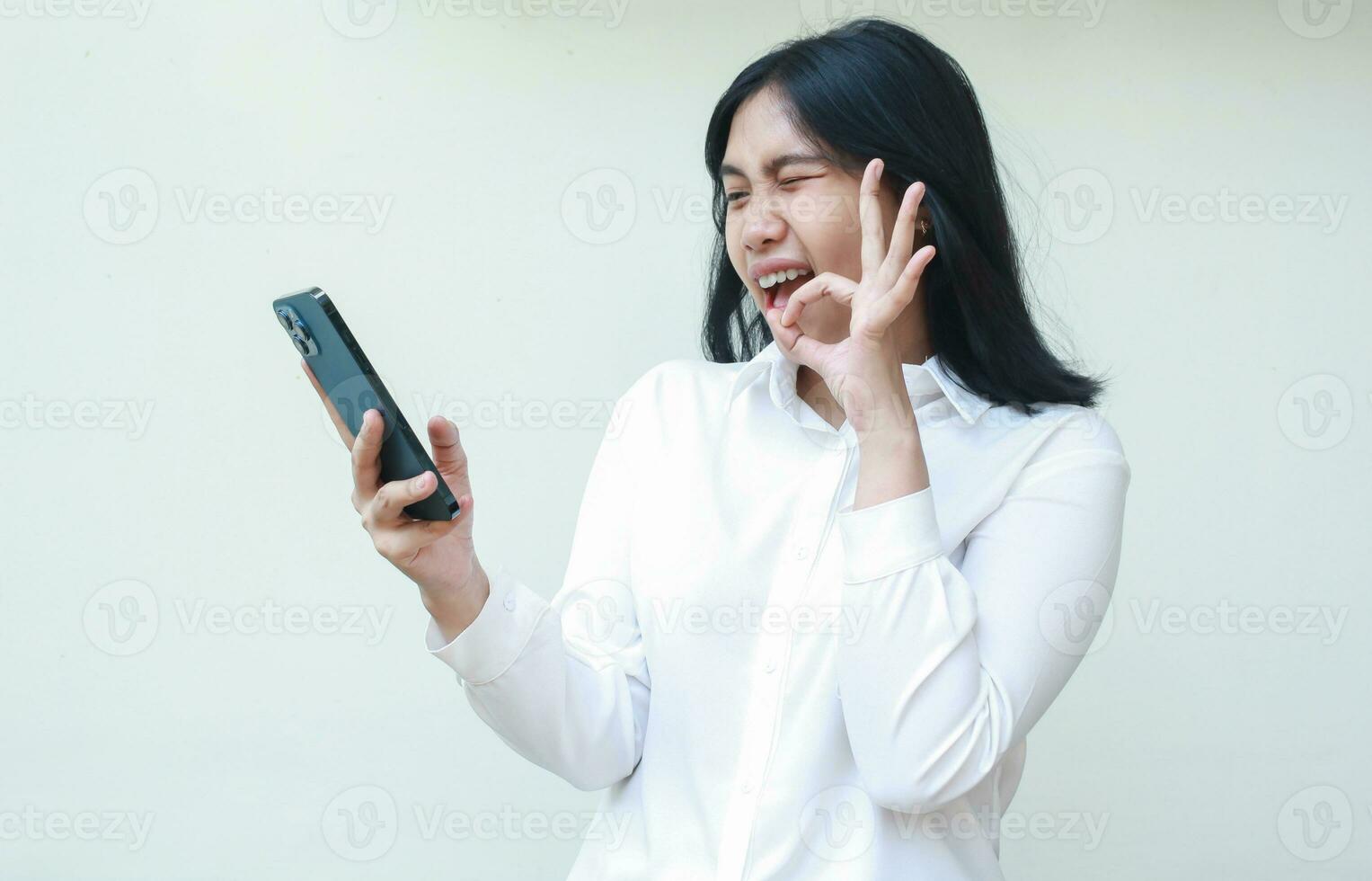 delighted asian young woman secretary holding mobile phone showing ok sign, okay symbol, wearing white formal suit shirt, flirting blink eye to screen in video call, agree gesture photo