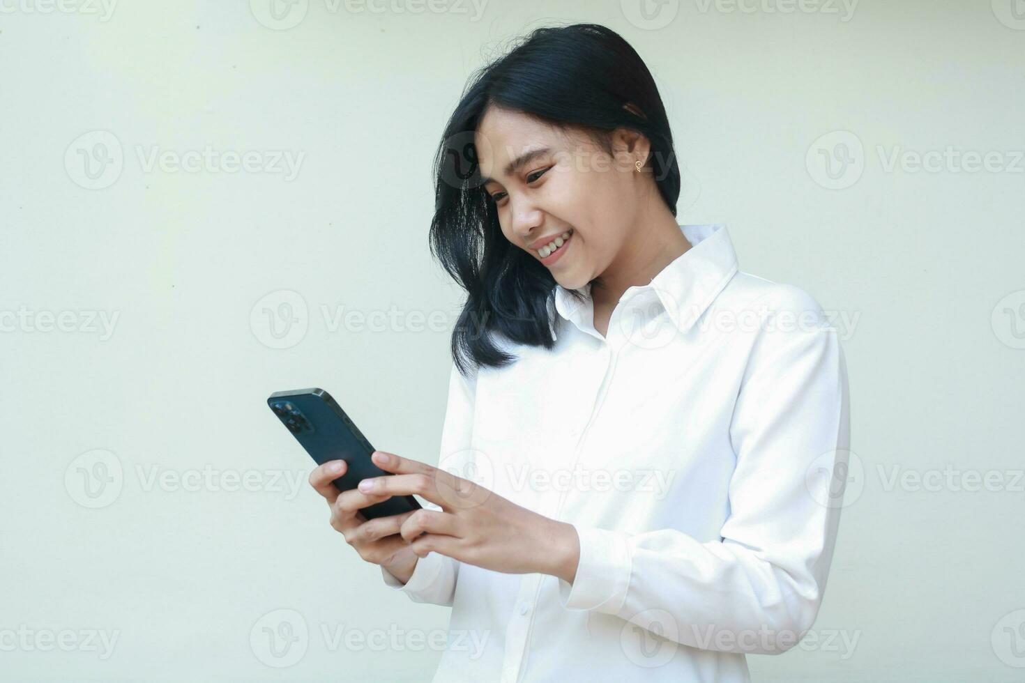 portrait of smiling asian young saleswoman holding smartphone surfing on social media, chatting message, read good news, wearing white formal suit shirt standing over isolated background photo