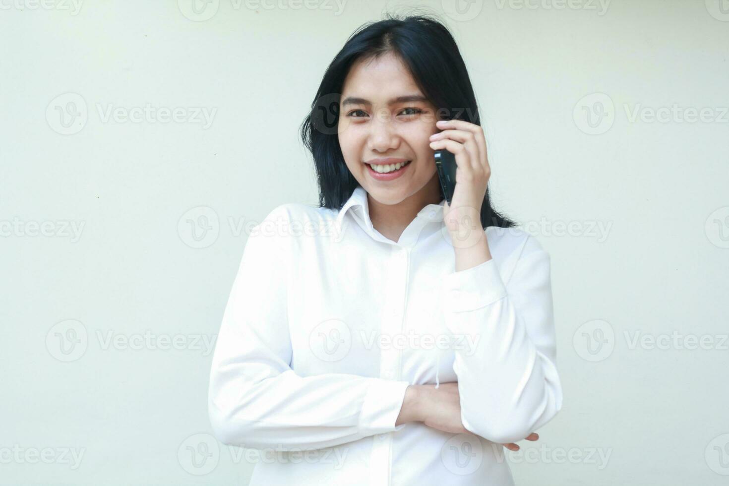 portrait of confident young asian business woman speaking on smartphone with arms folded looking at camera smiling peaceful wearing white formal suit shirt, standing isolated, wireless conversation photo