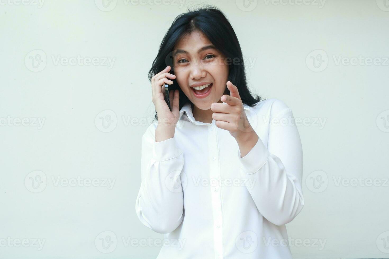 imagen de juguetón asiático joven vendedora Hablando en teléfono inteligente señalando dedo hacia adelante emocionado mirando a cámara espectáculo confidente sonrisa abierto boca vistiendo blanco formal camisa, sus usted gesto, en pie foto