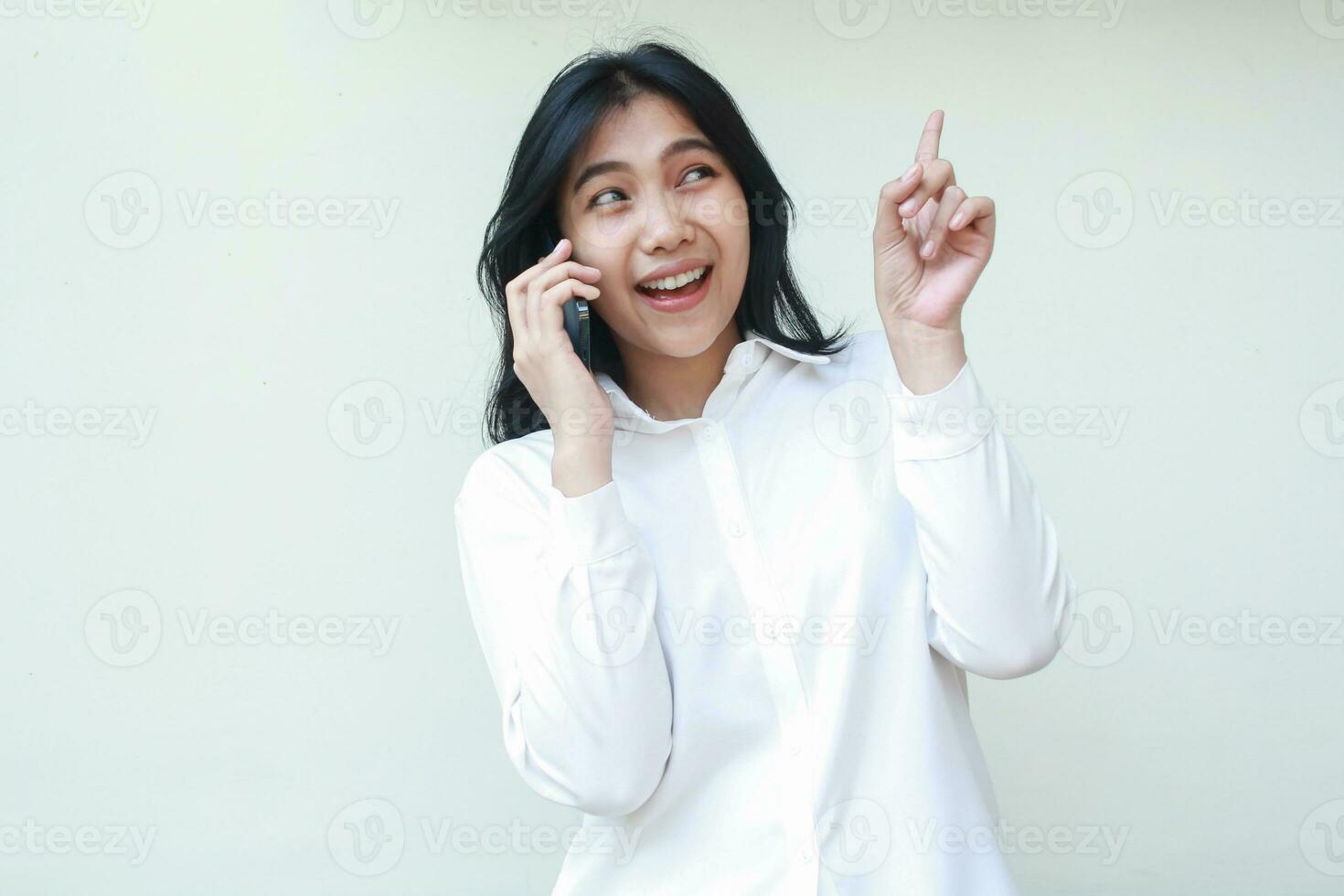 portrait pleased asian young woman secretary pointing finger up looking above to empty space while speaking on mobile phone wearing white formal suit shirt, wireless conversation, standing isolated photo