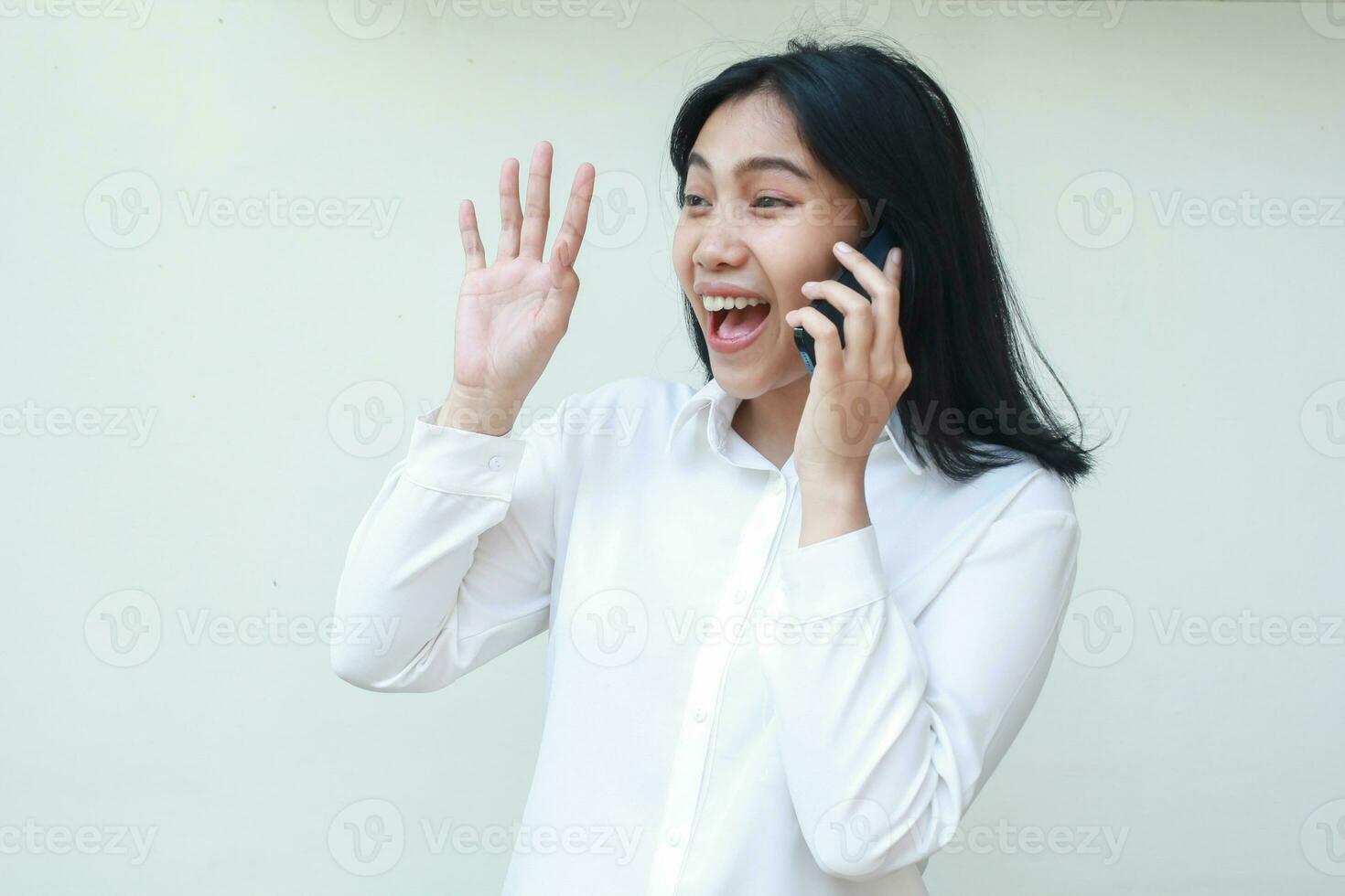 playful friendly asian business woman raising palms say hi and look away while talking on mobile phone wearing white suit formal shirt, said hello welcoming gesture, standing over isolated background photo