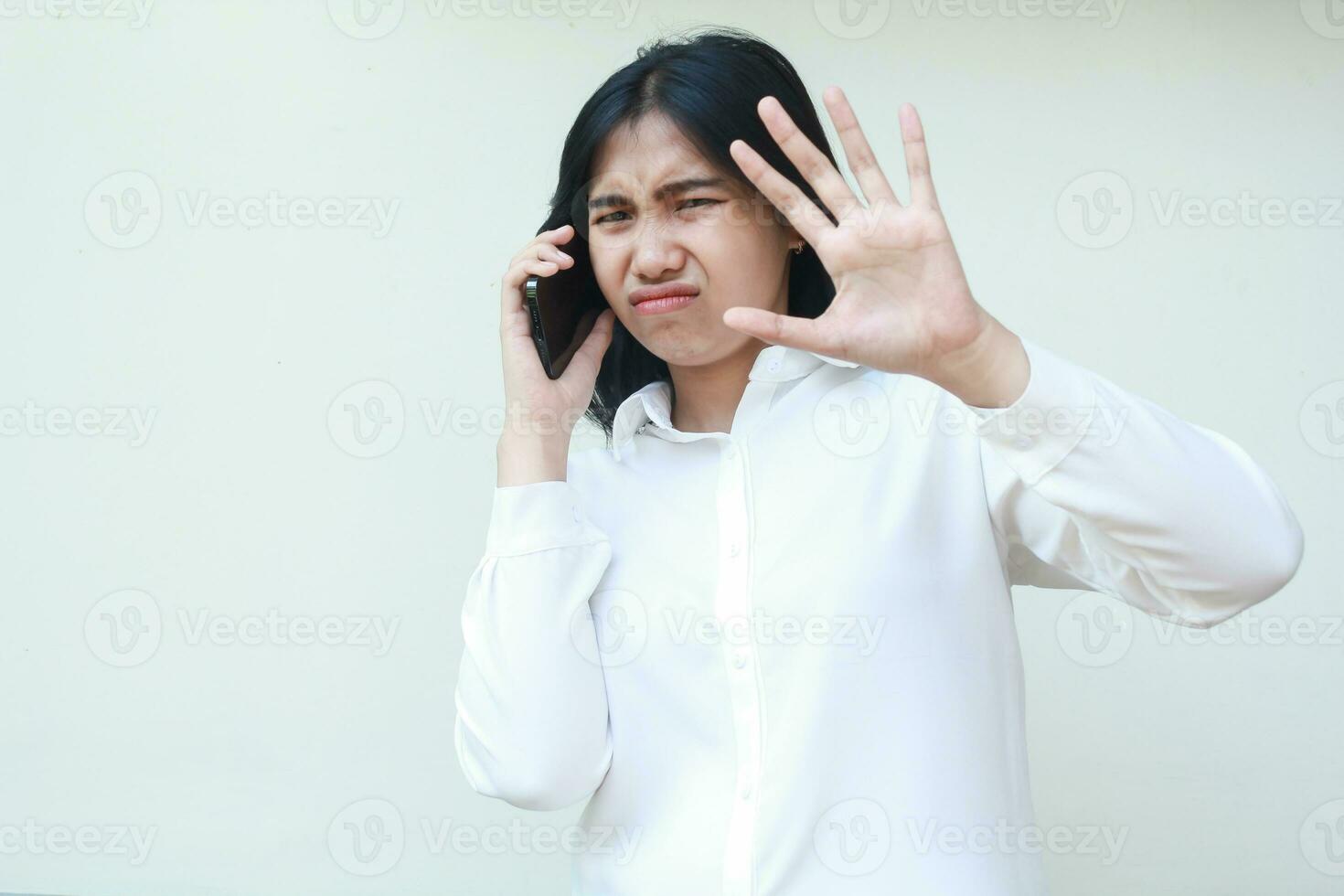 retrato de irritado asiático negocio mujer espectáculo triste expresión mientras Hablando en teléfono inteligente con levantamiento palmas demostración detener gesto vistiendo blanco camisa formal traje, en pie aislado foto