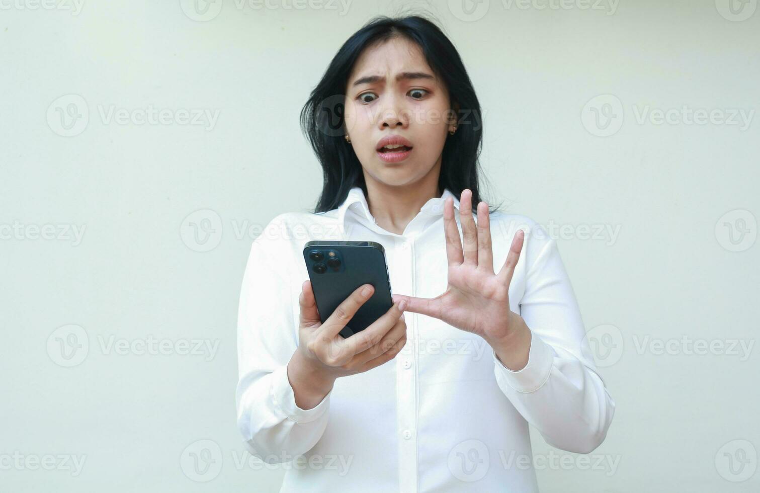 portrait of shocked asian business woman frightened looking at smartphone with raising palms showing stop gesture wearing white suit formal shirt, standing isolated, technology lifestyle concept photo