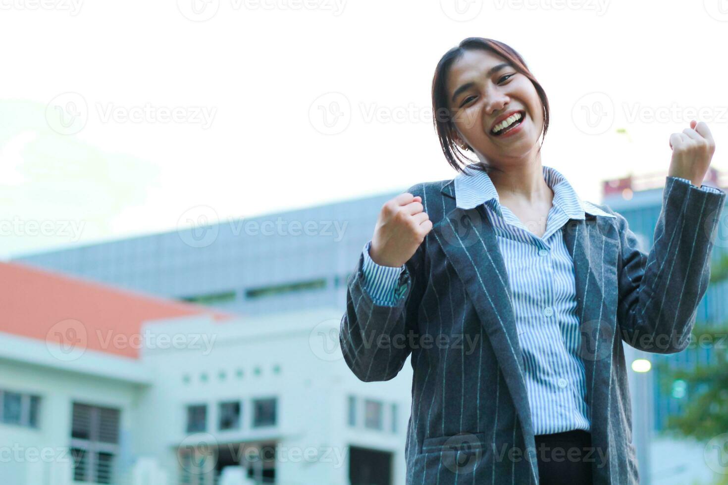 smiling asian business woman standing in city streets raising palms celebrate victory, yes gesture, wearing formal suit jacket, female executive standing in outdoors with urban building background photo