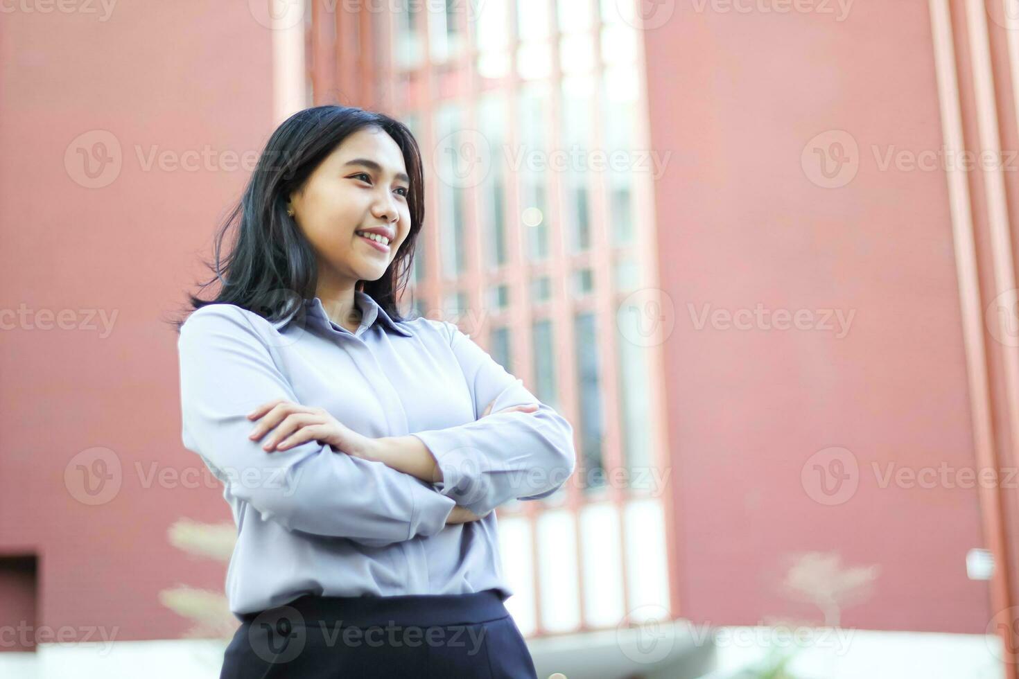 imagen de exitoso asiático negocio mujer en pie terminado oficina edificio con cruzar mano vistiendo formal camisa, hembra empresario sonriente confidente mirando lejos espectáculo optimista expresión foto